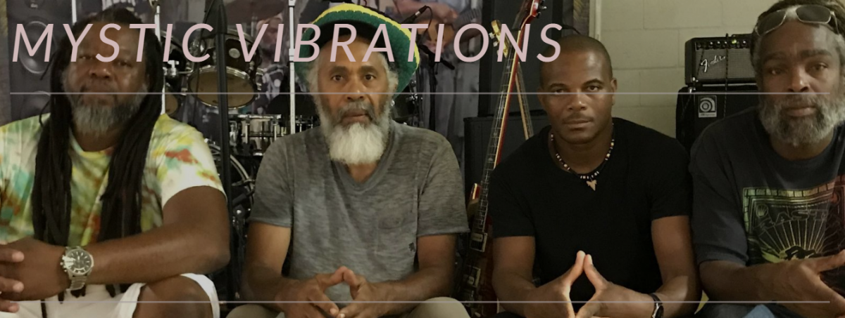 This is a picture of four African American men in the reggae band Mystic Vibrations sitting on the edge of a stage with musical equipment behind them in the background.