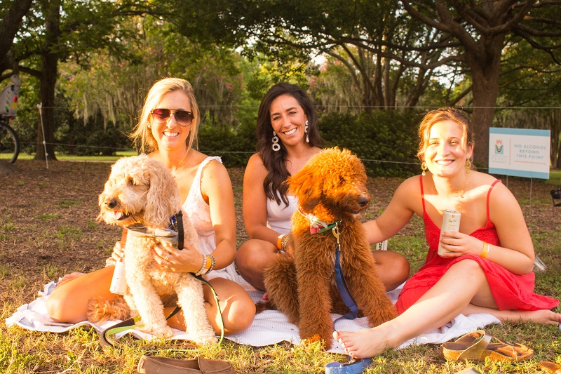 This is a picture of three women sitting on the ground drinking wine with a dog.